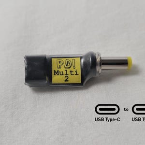 May include: A black PD! Multi 2 adapter with a silver and yellow tip. The adapter converts USB Type-C to USB Type-C. The adapter is on a white surface.