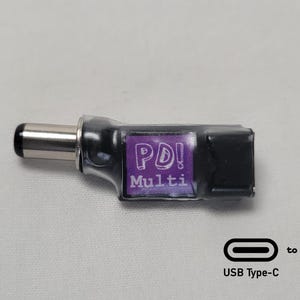 May include: A black PD Multi adapter with a silver tip and a purple label with white text. The adapter converts from USB Type-C to USB Type-C. The image shows the adapter on a white surface.