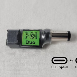 May include: A black and silver USB Type-C to USB Type-C adapter with a green label that reads "PD! Duo". The adapter is shown on a white surface, with USB Type-C symbols.