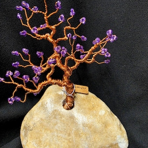 May include: A decorative wire tree sculpture featuring twisted copper-colored wire branches adorned with small purple amethyst beads. The tree is anchored into a smooth, light tan natural stone base and includes a small rectangular tag.
