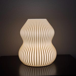 May include: A modern, decorative lamp with a unique hourglass shape. The lamp is made of vertical wooden slats that allow light to filter through. The lamp is lit from within, creating a warm, inviting glow. The lamp sits on a dark surface.