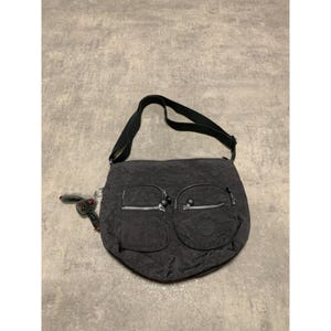 Op de afbeelding: Een donkergrijze schoudertas met twee ritsvakken aan de voorkant en een zwarte schouderriem. De tas heeft een kleine, bevestigde sleutelhanger met een aapachtig bedeltje. De tas is gemaakt van een textuurrijk materiaal.