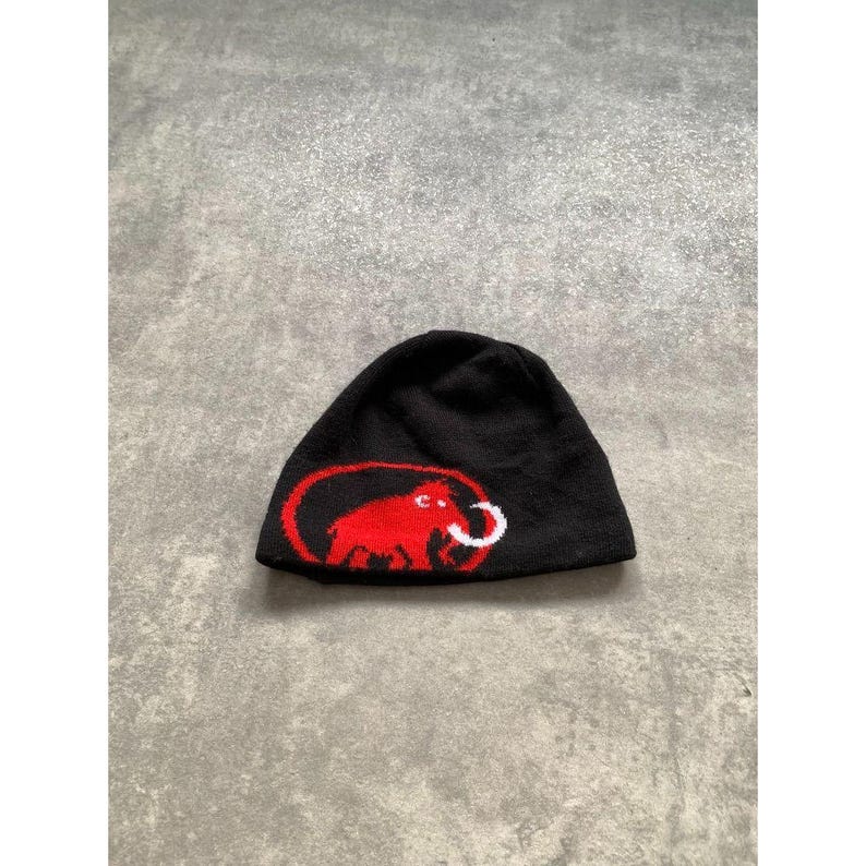 Op de afbeelding: Zwarte muts met een rood wollen mammoetlogo. Het logo staat in een rode ovaal met een witte slagtand. De muts is gemaakt van een zacht, gebreid materiaal, ideaal voor koud weer.