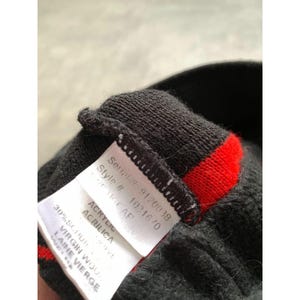 Op de afbeelding: Close-up van een zwarte gebreide muts met een rode streep. Een wit label toont het serienummer, het stijlnummer en de materiaalsamenstelling, inclusief acryl en scheerwol. De muts is een winteraccessoire.