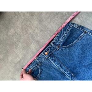 Pode incluir: Close-up de jeans azuis com uma fita m&eacute;trica rosa. Os jeans t&ecirc;m fecho de bot&atilde;o e bolsos. A fita m&eacute;trica &eacute; segurada ao longo da cintura, mostrando a medida em cent&iacute;metros.