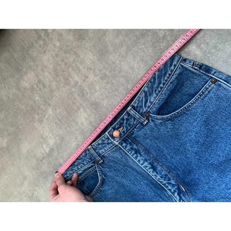 Pode incluir: Close-up de jeans azuis com uma fita m&eacute;trica rosa. Os jeans t&ecirc;m fecho de bot&atilde;o e bolsos. A fita m&eacute;trica &eacute; segurada ao longo da cintura, mostrando a medida em cent&iacute;metros.