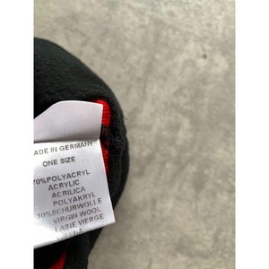 Op de afbeelding: Een zwarte gebreide muts met een rode binnenkant. Een wit label is bevestigd met de tekst "MADE IN GERMANY", "ONE SIZE" en de materiaalsamenstelling: 70% Polyacryl, 30% Scheerwol.