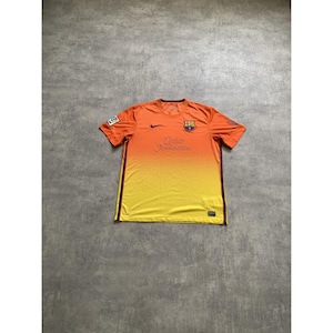 Op de afbeelding: Een oranje en geel voetbalshirt met korte mouwen en een kleurverloop, voorzien van het FC Barcelona-embleem op de linkerborst, een zwarte Nike-swoosh rechts, de tekst Qatar Foundation in het midden en een La Liga-patch op de mouw.