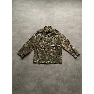 Könnte beinhalten: Eine Tarnjacke in Grün-, Braun- und Beigetönen. Die Jacke hat eine Knopfleiste, zwei Brusttaschen und zwei untere Taschen. Sie hat lange Ärmel und einen klassischen Kragen. Das Kleidungsstück liegt flach auf einer grauen Oberfläche.