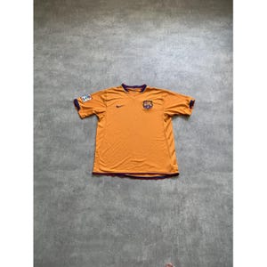 Op de afbeelding: Een oranje voetbalshirt met korte mouwen en een V-hals met donkerblauwe en rode biezen. Het shirt is voorzien van een zwart Nike-logo op de rechterborst en het clubembleem van FC Barcelona op de linkerborst, met een LFP-competitiepatch op de mouw.