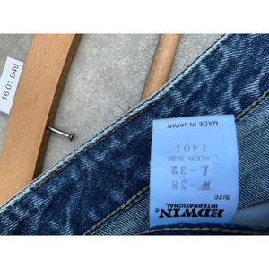Pode incluir: Close-up de cal&ccedil;as de ganga azuis com uma etiqueta que diz "EDWIN INTERNATIONAL" e "MADE IN JAPAN". A etiqueta tamb&eacute;m indica o tamanho W-38 e L-32. Um cabide de madeira &eacute; parcialmente vis&iacute;vel.