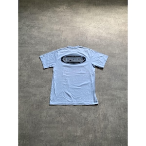 May include: A light blue short-sleeve t-shirt featuring a dark oval graphic on the upper back. The graphic contains the text RIPCURL in a bold, stylized font, framed by a dark border with small star accents on the left and right sides.