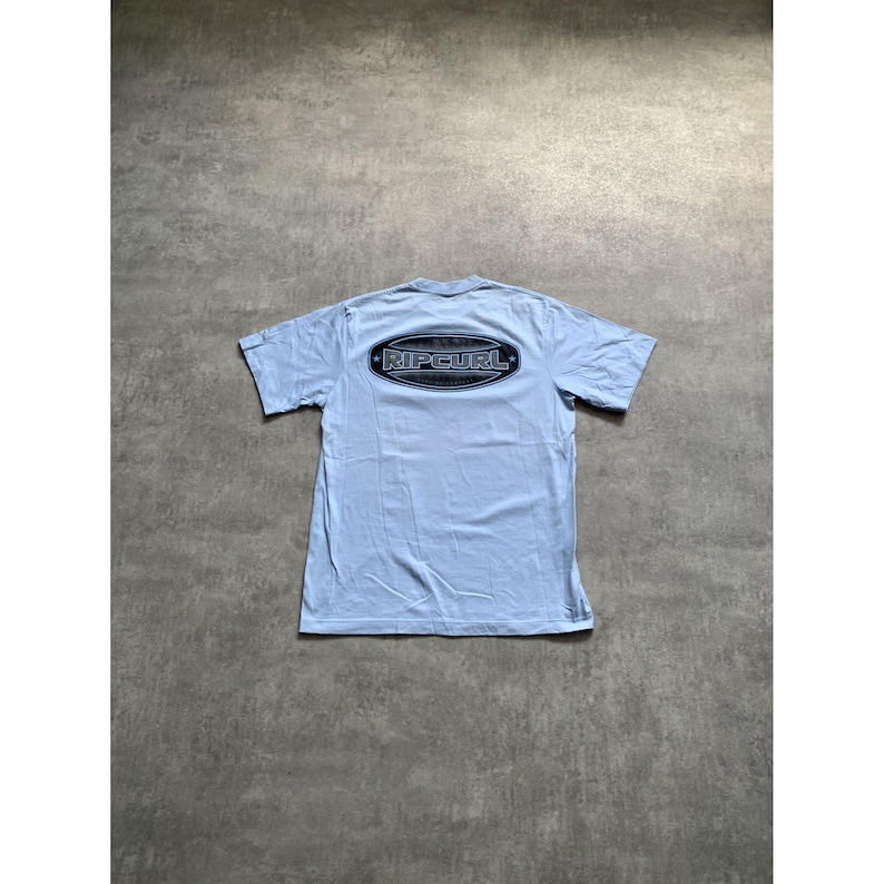 May include: A light blue short-sleeve t-shirt featuring a dark oval graphic on the upper back. The graphic contains the text RIPCURL in a bold, stylized font, framed by a dark border with small star accents on the left and right sides.