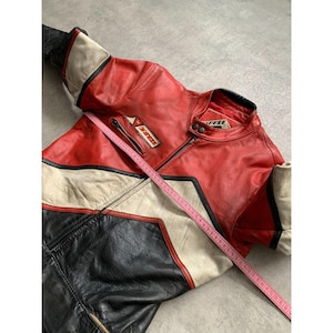 May include: Red, black, and white leather motorcycle jacket with the brand name "Dainese" visible. The jacket has a zippered front, a stand-up collar, and a pocket on the chest. A pink measuring tape is laid across the jacket.