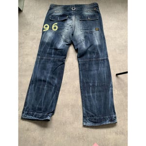 May include: Blue denim jeans with a faded wash and a relaxed fit. The jeans feature a yellow "96" graphic on the back pocket. They have multiple pockets, a belt loop, and a button and zipper closure. The jeans are laid flat on a light gray surface.