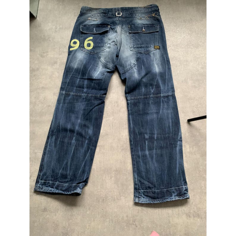 May include: Blue denim jeans with a faded wash and a relaxed fit. The jeans feature a yellow "96" graphic on the back pocket. They have multiple pockets, a belt loop, and a button and zipper closure. The jeans are laid flat on a light gray surface.