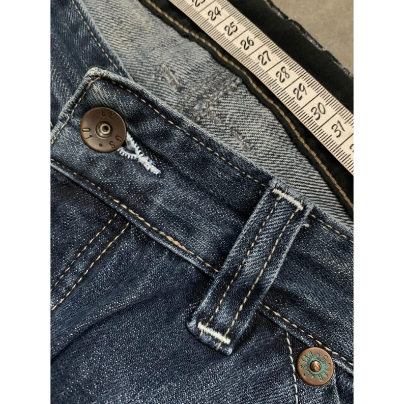 May include: Close-up of blue denim jeans with a button and belt loop. The jeans have white stitching and a metal button. A measuring tape is placed across the jeans, showing measurements in inches.