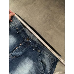 May include: Close-up of blue denim jeans with button closure and pockets. A measuring tape is placed along the waistband, showing the waist measurement. The jeans have a faded wash and a classic five-pocket design.
