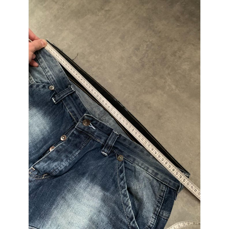 May include: Close-up of blue denim jeans with button closure and pockets. A measuring tape is placed along the waistband, showing the waist measurement. The jeans have a faded wash and a classic five-pocket design.