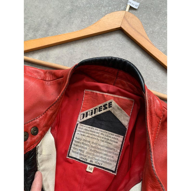 May include: Red leather motorcycle jacket with black and white accents. The jacket features a black collar and a label with the brand name "DAINESE" and the text "MADE IN ITALY" and size 50. The jacket is hanging on a wooden hanger.