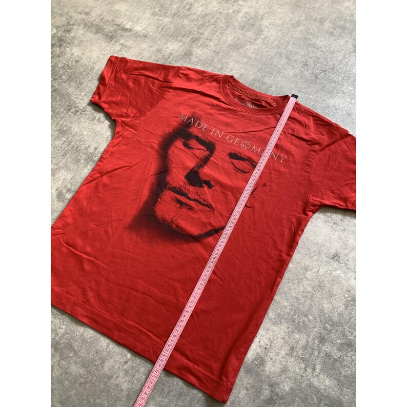 Peut inclure: Un t-shirt rouge &agrave; manches courtes avec un graphique sombre repr&eacute;sentant un visage et le texte "MADE IN GERMANY". Un ruban &agrave; mesurer rose est plac&eacute; en diagonale sur le t-shirt, indiquant une longueur d'environ 69 cm depuis le col.