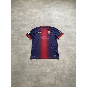 Op de afbeelding: Een marineblauw en rood voetbalshirt met korte mouwen, voorzien van een geel Nike-logo op de rechterborst, het FC Barcelona-embleem op de linkerborst en de tekst Qatar Foundation in het geel gedrukt in het midden van de borst.