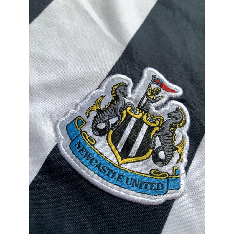 May include: A black and white striped jersey featuring the embroidered Newcastle United football club crest. The emblem displays two seahorses flanking a central shield with vertical stripes, topped by a small flag, all above a blue banner.