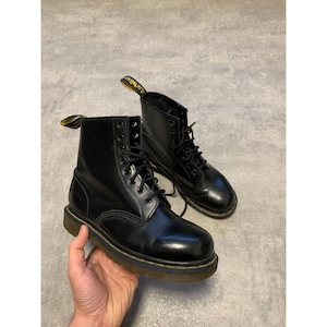 Puede incluir: Un par de botas de cuero negro con un acabado brillante. Las botas tienen un diseño con cordones, costuras amarillas y una lengüeta amarilla. Las botas miden aproximadamente 15 cm de alto.