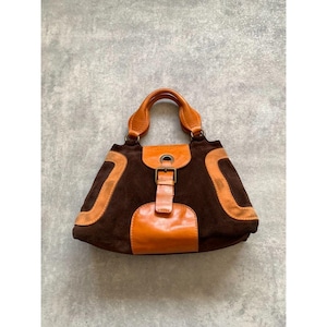 May include: A brown suede handbag with orange leather accents. The bag features a top handle, a buckle closure, and orange leather trim. The bag's design includes a curved shape and a structured silhouette.