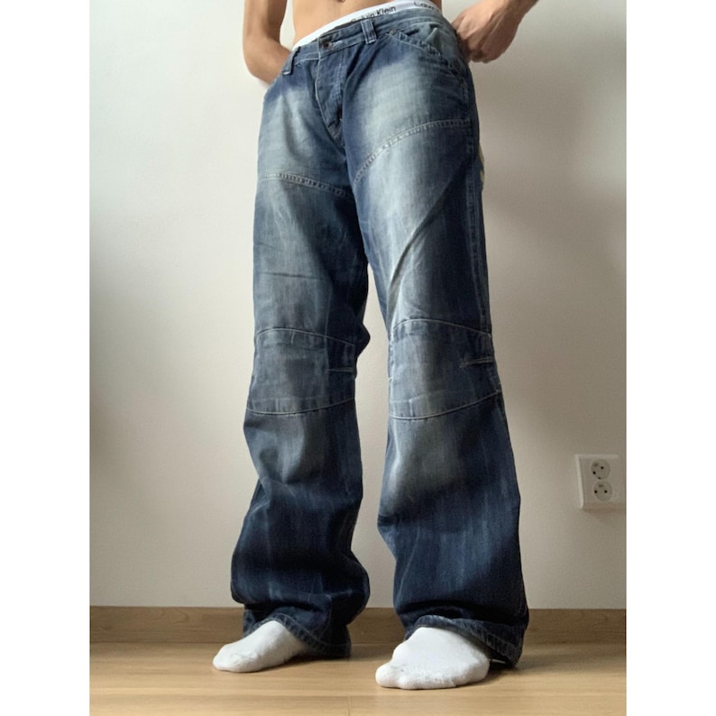 May include: Blue denim jeans with a relaxed fit. The jeans have a faded wash and feature reinforced panels on the knees. The jeans have a button and zipper closure. The jeans are worn with white socks.