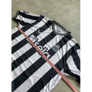 May include: A black and white striped soccer jersey featuring the Newcastle United crest, the Castor logo, and the Sela sponsor text. A pink measuring tape is placed diagonally across the front, showing measurements in inches for sizing reference.