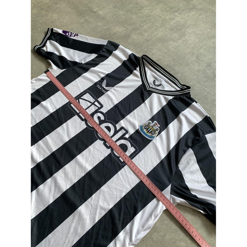 May include: A black and white striped soccer jersey featuring the Newcastle United crest, the Castor logo, and the Sela sponsor text. A pink measuring tape is placed diagonally across the front, showing measurements in inches for sizing reference.