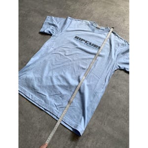 May include: A light blue short-sleeve t-shirt featuring the text RIPCURL in black lettering across the chest. A white measuring tape is placed diagonally across the shirt, showing a length measurement of approximately 29 inches from the shoulder.