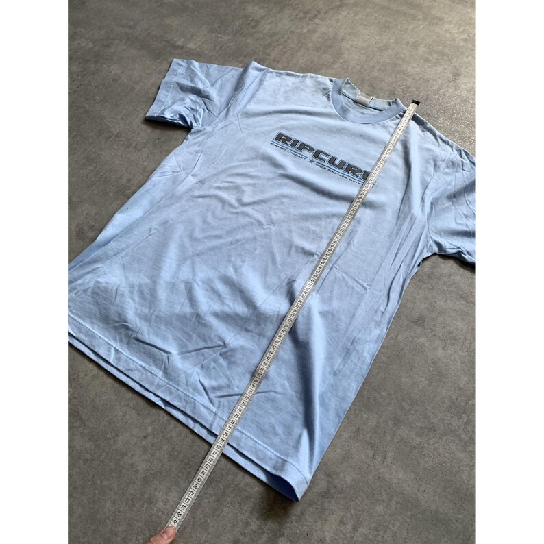 May include: A light blue short-sleeve t-shirt featuring the text RIPCURL in black lettering across the chest. A white measuring tape is placed diagonally across the shirt, showing a length measurement of approximately 29 inches from the shoulder.