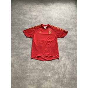 May include: A red short-sleeved athletic jersey featuring gold stripes along the shoulders, a gold Adidas logo on the right chest, and a gold Spanish national football team crest on the left chest. The shirt has a crew neckline and a curved hem.