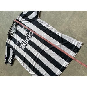 May include: A black and white striped short-sleeve jersey with a V-neck collar, featuring the text 'sela' and a team crest on the chest. A pink measuring tape is placed diagonally across the fabric, showing a length of approximately 30 inches.
