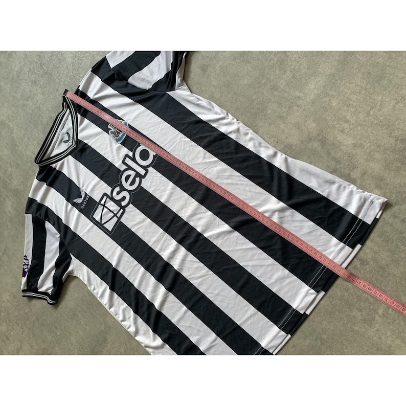 May include: A black and white striped short-sleeve jersey with a V-neck collar, featuring the text 'sela' and a team crest on the chest. A pink measuring tape is placed diagonally across the fabric, showing a length of approximately 30 inches.