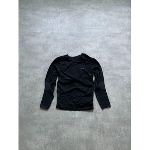 Puede incluir: Camiseta negra de manga larga con cuello en V. La camiseta tiene un pequeño logotipo en el pecho y coderas. La tela parece ser un material ligero y deportivo.