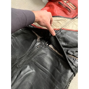 May include: Black leather motorcycle jacket with red and cream-colored accents. The jacket features multiple zippers and a logo patch on the upper right shoulder. The jacket appears to be well-worn and has a classic design.