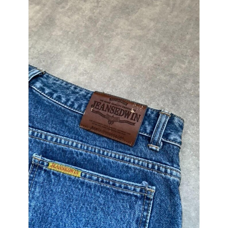 Pode incluir: Close-up de cal&ccedil;as de ganga azuis com uma etiqueta de couro castanho JEANS EDWIN. A etiqueta tem texto em relevo e um log&oacute;tipo. As cal&ccedil;as de ganga t&ecirc;m uma etiqueta amarela JEANS EDWIN no bolso de tr&aacute;s.