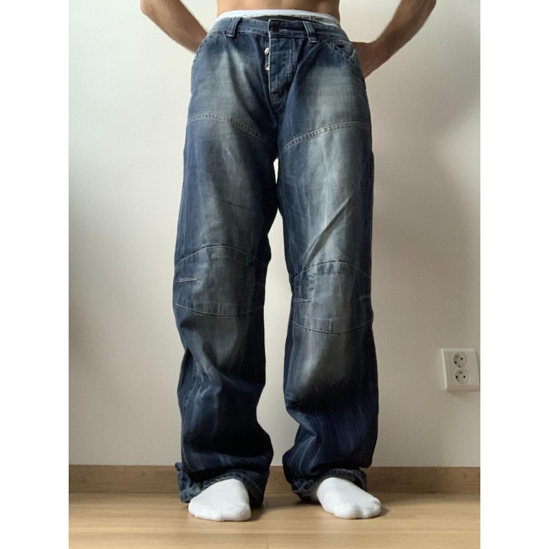 May include: Dark wash denim jeans with a relaxed fit. The jeans feature a button fly and reinforced knee panels. The denim has a faded, worn look. The jeans are paired with white socks.
