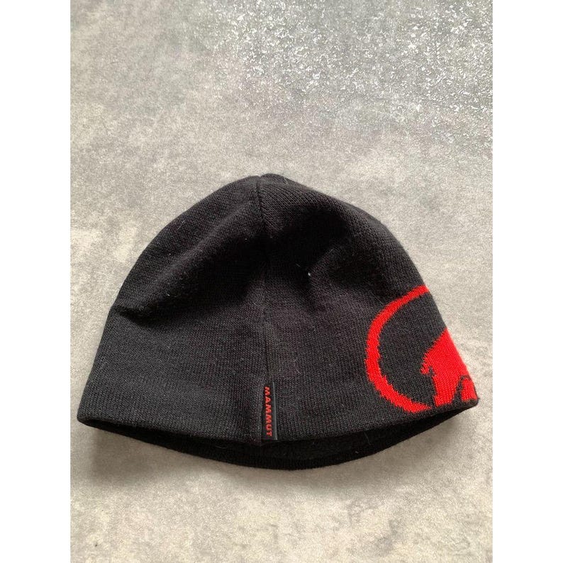 Op de afbeelding: Zwarte gebreide beanie met een rood logo aan de zijkant. De beanie heeft de merknaam "MAMMUT" in het rood. De hoed is gemaakt van een zacht, gebreid materiaal.