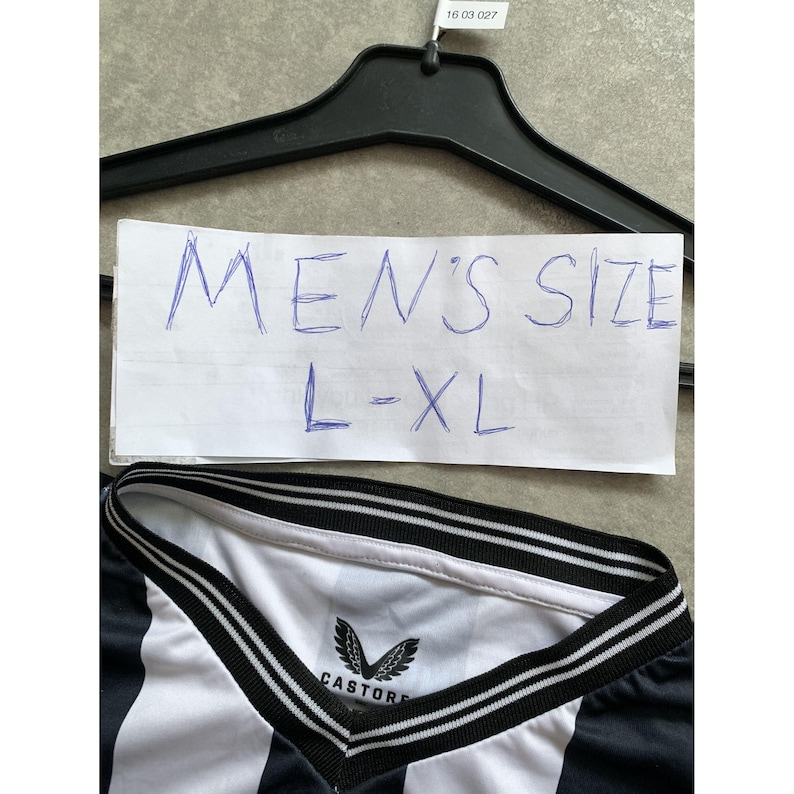 May include: A black and white striped shirt with a V-neck collar featuring black and white trim. The interior neck label displays the Castore brand logo and name. A white paper sign above the shirt displays the handwritten text MEN'S SIZE L-XL.