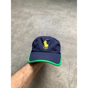 Op de afbeelding: Marineblauwe baseballcap met een geel logo van een polospeler. De rand is groen omlijnd. De cap is gemaakt van een lichtgewicht materiaal.