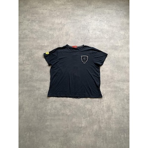 May include: A black short-sleeved t-shirt featuring a small yellow Ferrari logo on the right sleeve and a larger, outlined Ferrari shield emblem printed in white on the left chest area. The shirt has a standard crew neckline and a red interior neck label.