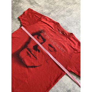Peut inclure: Un t-shirt rouge &agrave; manches courtes avec un graphique noir repr&eacute;sentant un visage et le texte MADE IN GERMANY. Un ruban &agrave; mesurer rose est plac&eacute; en diagonale sur la poitrine, indiquant une largeur d'environ 56 cm sur le tissu.