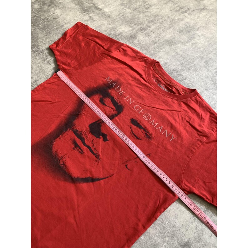 Peut inclure: Un t-shirt rouge &agrave; manches courtes avec un graphique noir repr&eacute;sentant un visage et le texte MADE IN GERMANY. Un ruban &agrave; mesurer rose est plac&eacute; en diagonale sur la poitrine, indiquant une largeur d'environ 56 cm sur le tissu.