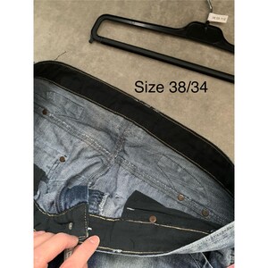 May include: Close-up of blue denim jeans with a black waistband and button closure. The jeans are size 38/34. A black hanger is visible in the background.