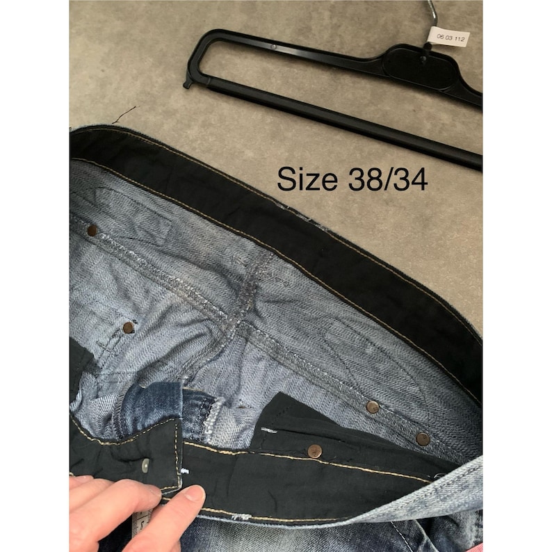 May include: Close-up of blue denim jeans with a black waistband and button closure. The jeans are size 38/34. A black hanger is visible in the background.