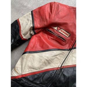 May include: A vintage red, black, and white leather motorcycle jacket. The jacket features a zippered pocket and a "DAINESE" patch. The jacket has a classic racing design with color-blocked panels and a worn appearance.